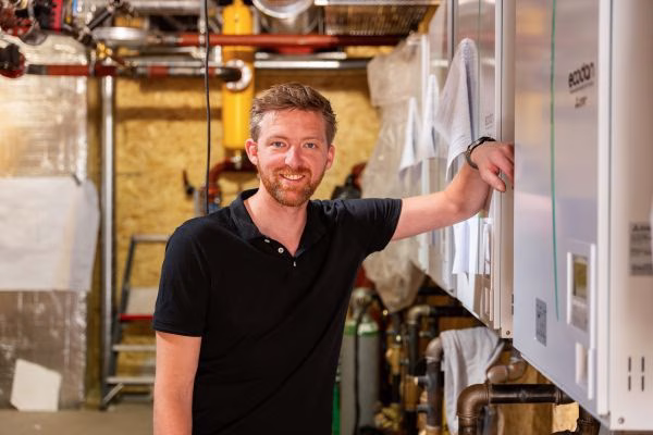 Man in een brouwerij naast brouwinstallaties, glimlachend en een kraan vasthoudend.
